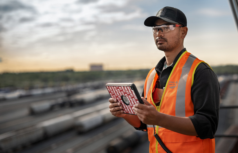 Ein Bahnmitarbeiter, umgeben von grossen Waggons, steht mit einem iPad in der Hand und schaut nach vorne