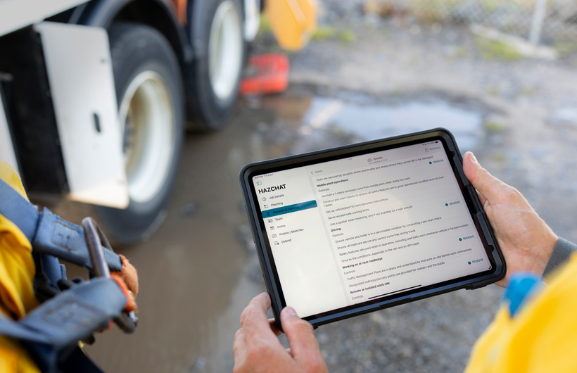 Zwei Hände halten ein iPad mit geöffneter HazChat App, die Jobdetails, Planung, Gefahren und Kontrollen anzeigt, während eine Person auf einer Baustelle neben einer Schlammpfütze steht